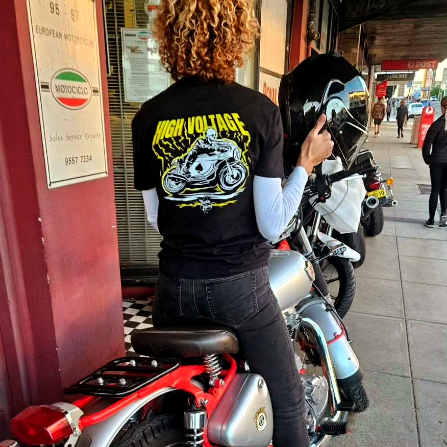 A black t-shirt with a high voltage design featuring a skeleton riding an old school Italian cafe racer motorcycle with electricity sparks, Genovieve wearing High Voltage t-shirt whilst visiting Motociclo in Newtown Sydney whilst on her Royal Enfield classic trials 500
